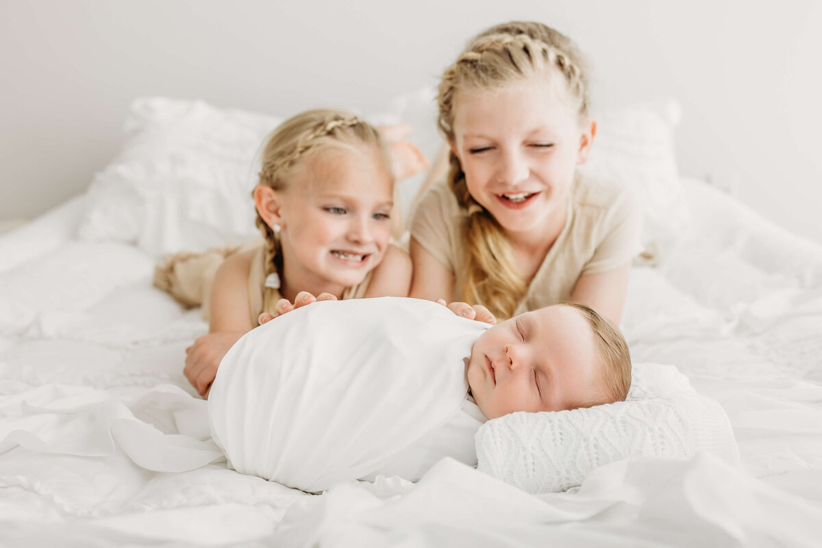 Happy sisters looking at sleeping new baby brother on all white bed
