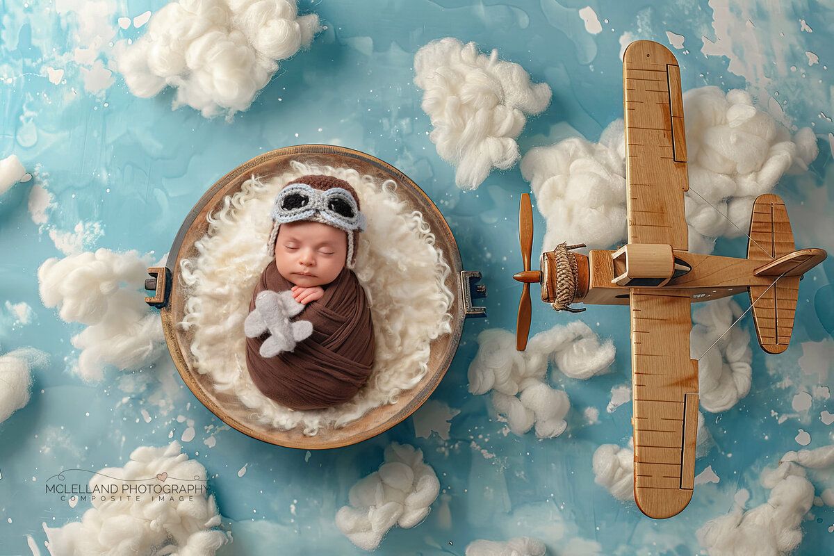 Newborn boy in an aviator-themed setup with fluffy clouds and a wooden airplane prop.