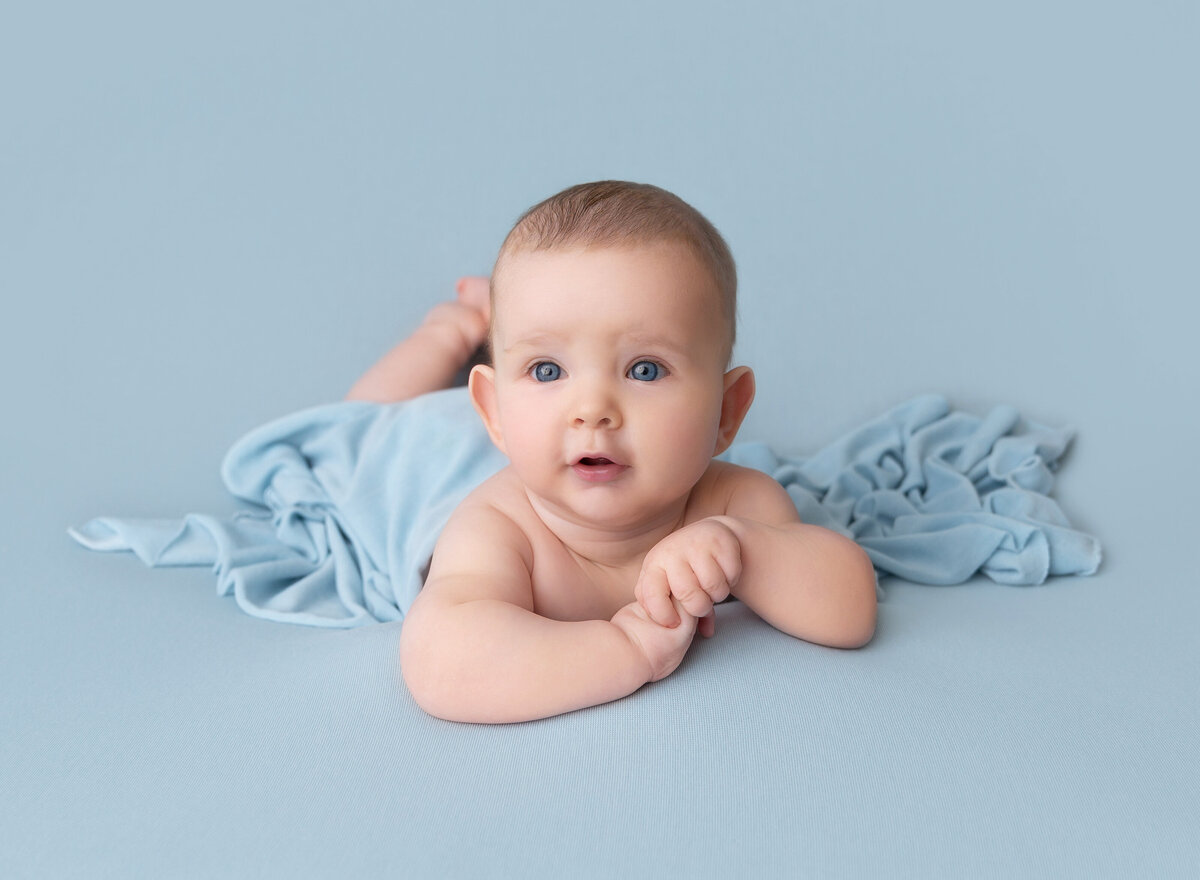 Baby boy is lying on his belly and propping his head up for a 6-month milestone photoshoot. Baby is looking at the camera with a smile.