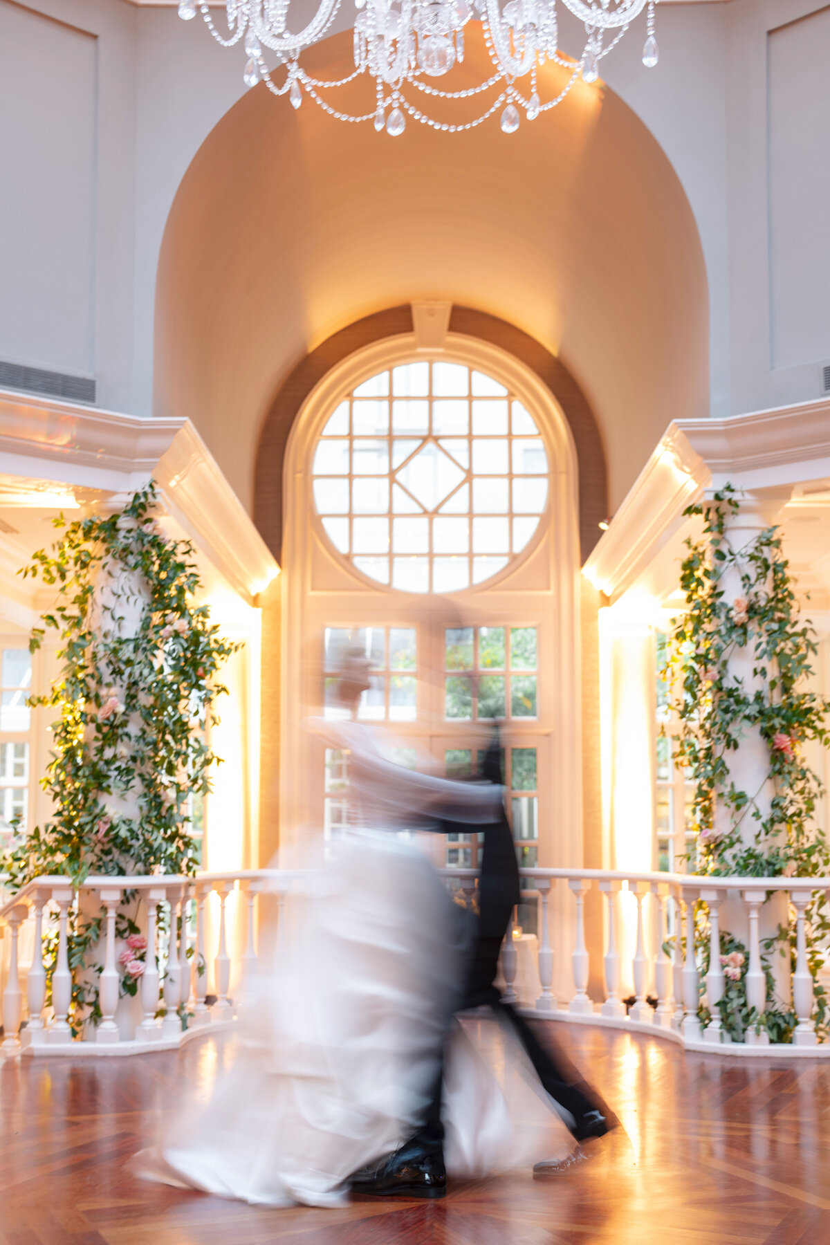 A blurred couple dances gracefully in an elegant, well-lit room with a large arched window and a chandelier. The walls are adorned with greenery, and the scene exudes a romantic and dreamy atmosphere.