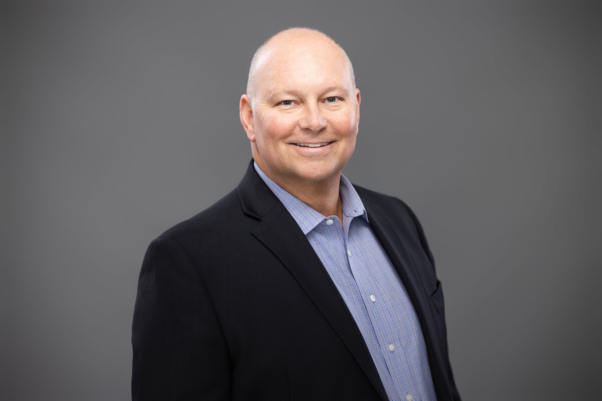 This professional portrait features Milton Snyder, confidently photographed against a textured backdrop in Cincinnati. The image exudes professionalism with Milton’s polished look in a classic black blazer over a blue checkered shirt. His warm smile and direct gaze create an inviting yet authoritative presence. The portrait is expertly lit to highlight his features and is perfect for corporate use, demonstrating a strong personal brand. He is bald but is very distinguished.