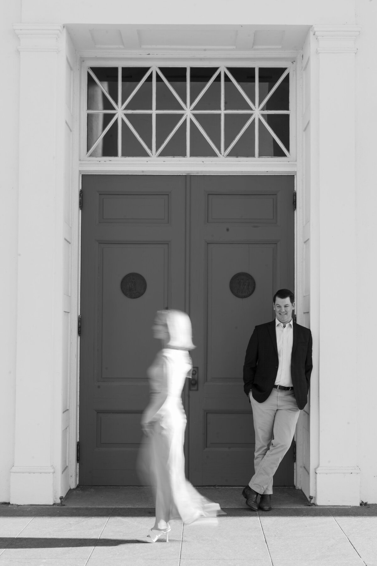 Richmond VA Engagement Photographer | blurry black and white image of girl walking past her fiancé who is leaning against the doorway of the Virginia Capitol Building in Richmond, Virginia