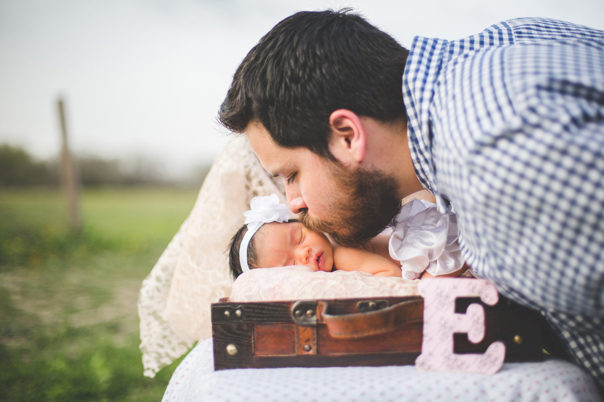 Father kissing his newborn daughter sleeping in a vintage suitcase in a field in San Antonio