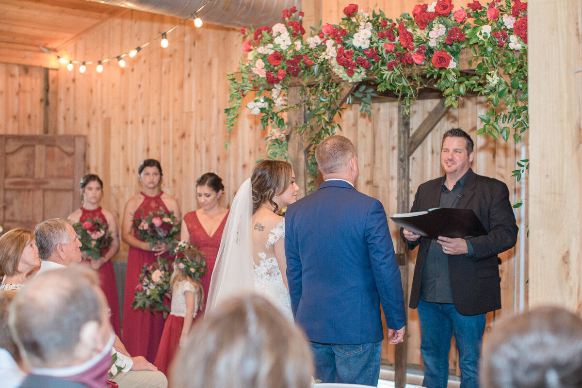 The bride and groom stand at the in front of their wedding arch, which is decorated with white and red roses