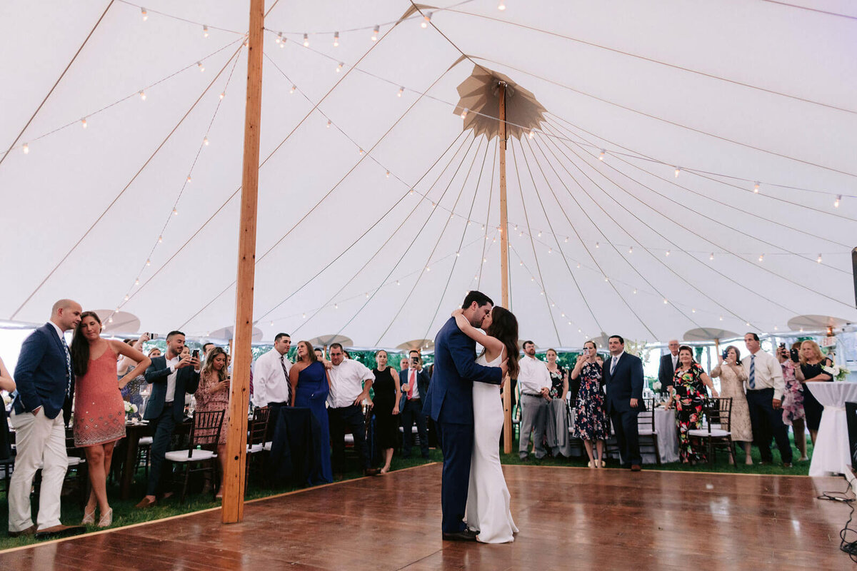The bride and groom are kissing on the dance floor, as the guests watch on, in Cape Cod Summer Tent, MA.