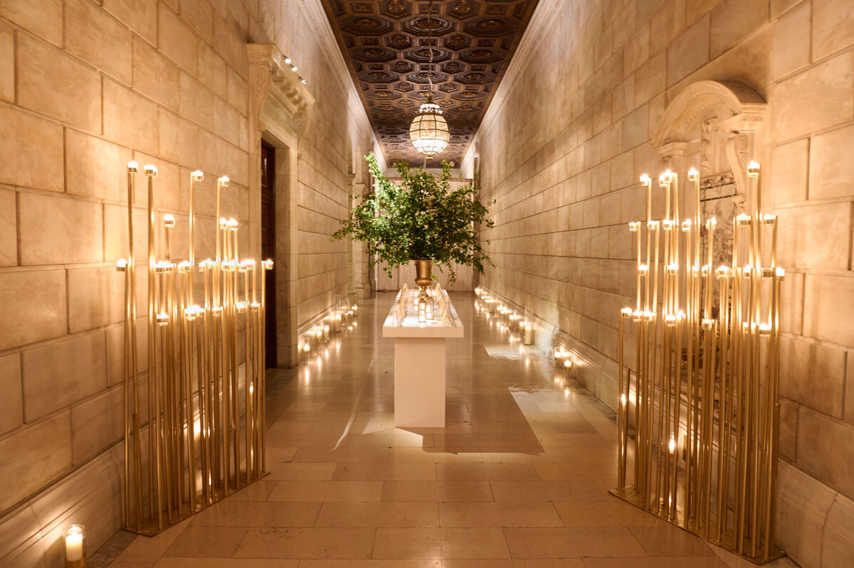 In the magnificent hallway with marble walls, lit by numerous candles mounted on tall, slender stands, located at The New York Public Library, photographed by the talented Shawn Connell. The ceiling features intricate wooden carvings and a chandelier that hangs majestically in the center. At the far end of the hallway, a white pedestal supports a large, lush arrangement of greenery, adding a touch of natural elegance to the grandeur of the space. Videography captured by Aaron Navak Films.
