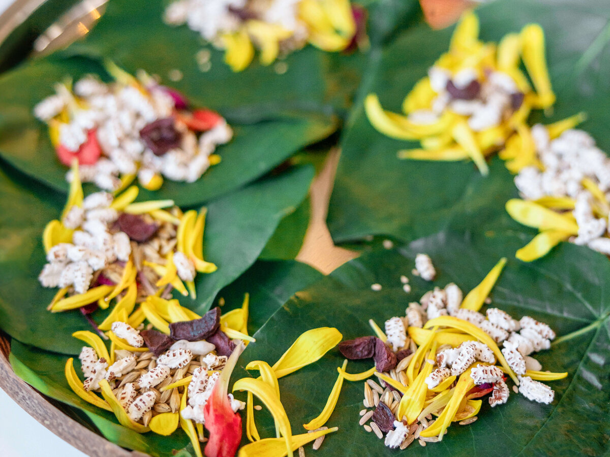A tray displays green leaves topped with a colorful arrangement of popped grains, yellow flower petals, red petals, and small seeds. The natural elements create a vibrant and decorative pattern.