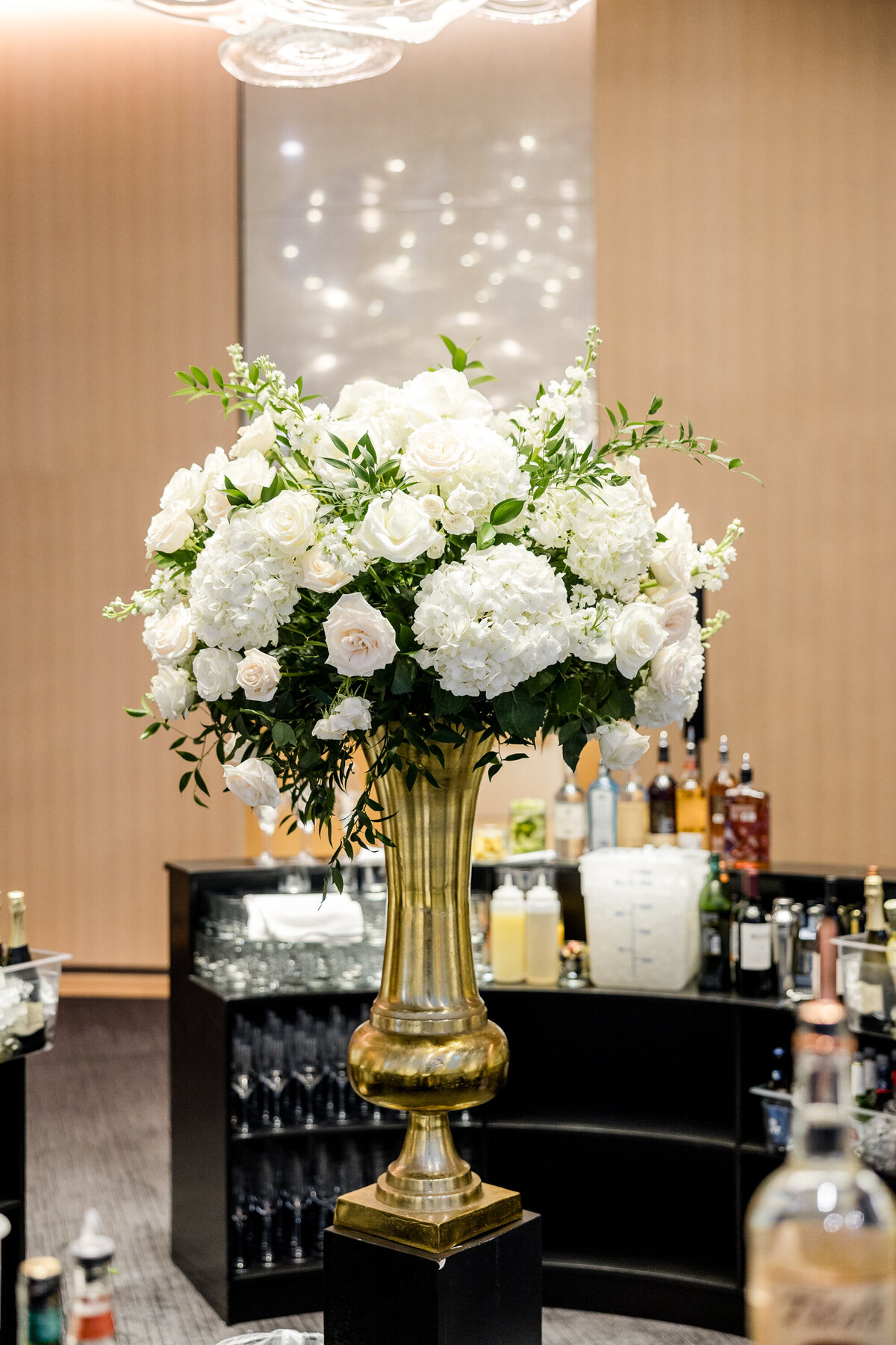 A grand white flower arrangement in a tall gold vase displayed on a bar, at The Conrad Washington DC Hotel adding elegance to the AAREP gala reception.