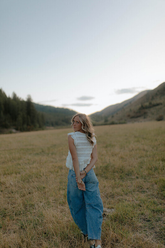 A girl looks over her shoulder at the camera for a senior photo. She's standing outside with trees and mountains behind her in the distance.