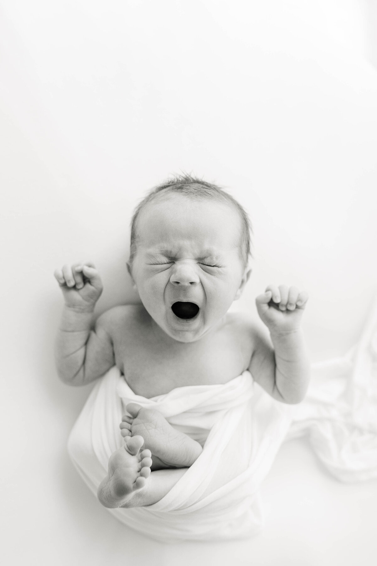 Black and white photo of a newborn baby yawning with his hands in little fists by his head and his torso wrapped in a white blanket with his feet out during a session with Indianapolis newborn photographer, Brittney Lear.