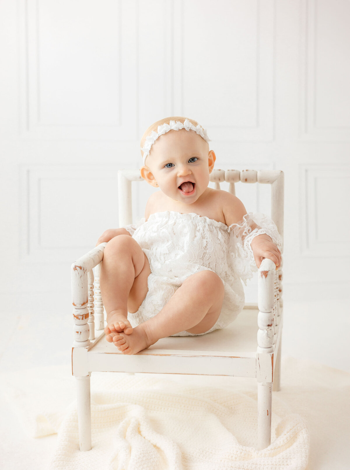 a little girl wearing a white lace romper sitting in a white chair smiling for her one year old pictures in Savannah, ga with Constance Calton Photography