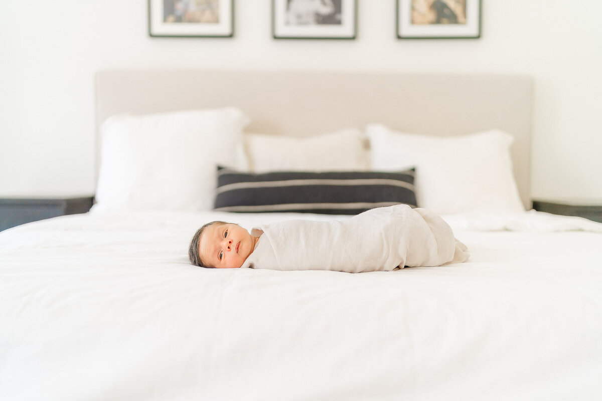 a newborn boy is swaddled in a white blanket and lays on a bed in his Round Rock home.