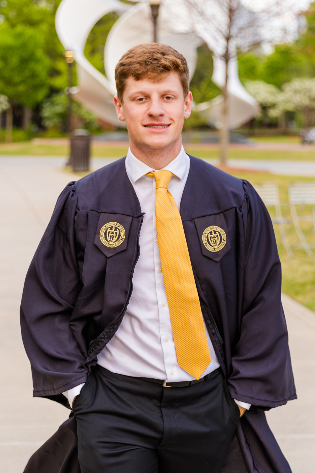 Undergrad senior boy walking on Georgia Tech campus hands in pockets wearing graduation gown over a white shirt and a yellow tie during graduation photo session with Atlanta photographer Laure Photography