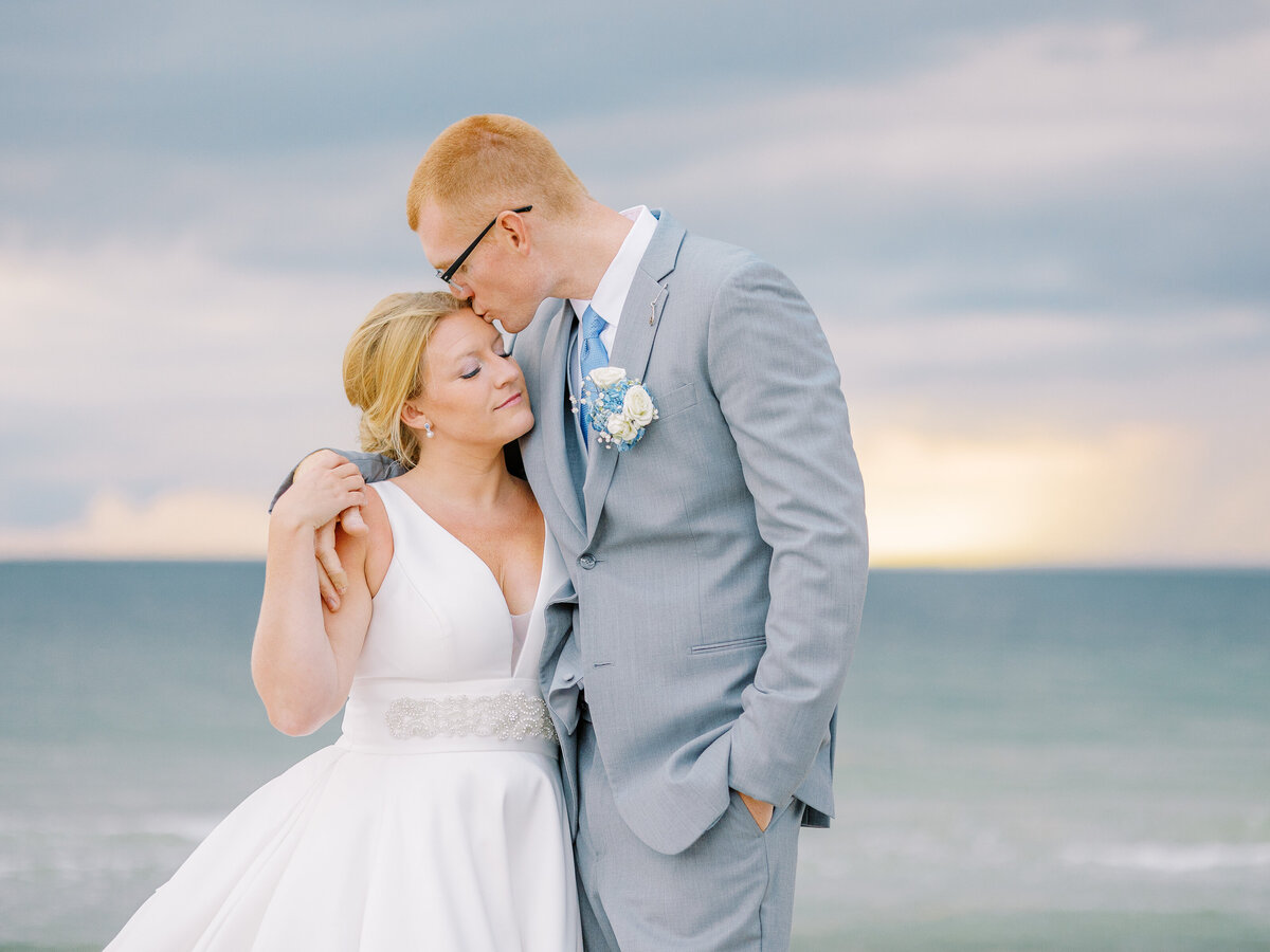 Groom kissing bride on the forehead in front of a sunset on the water