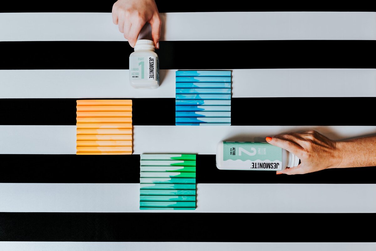 Black and white striped backdrop with multiple variously colored paint molds and two hands holding paint supplies.