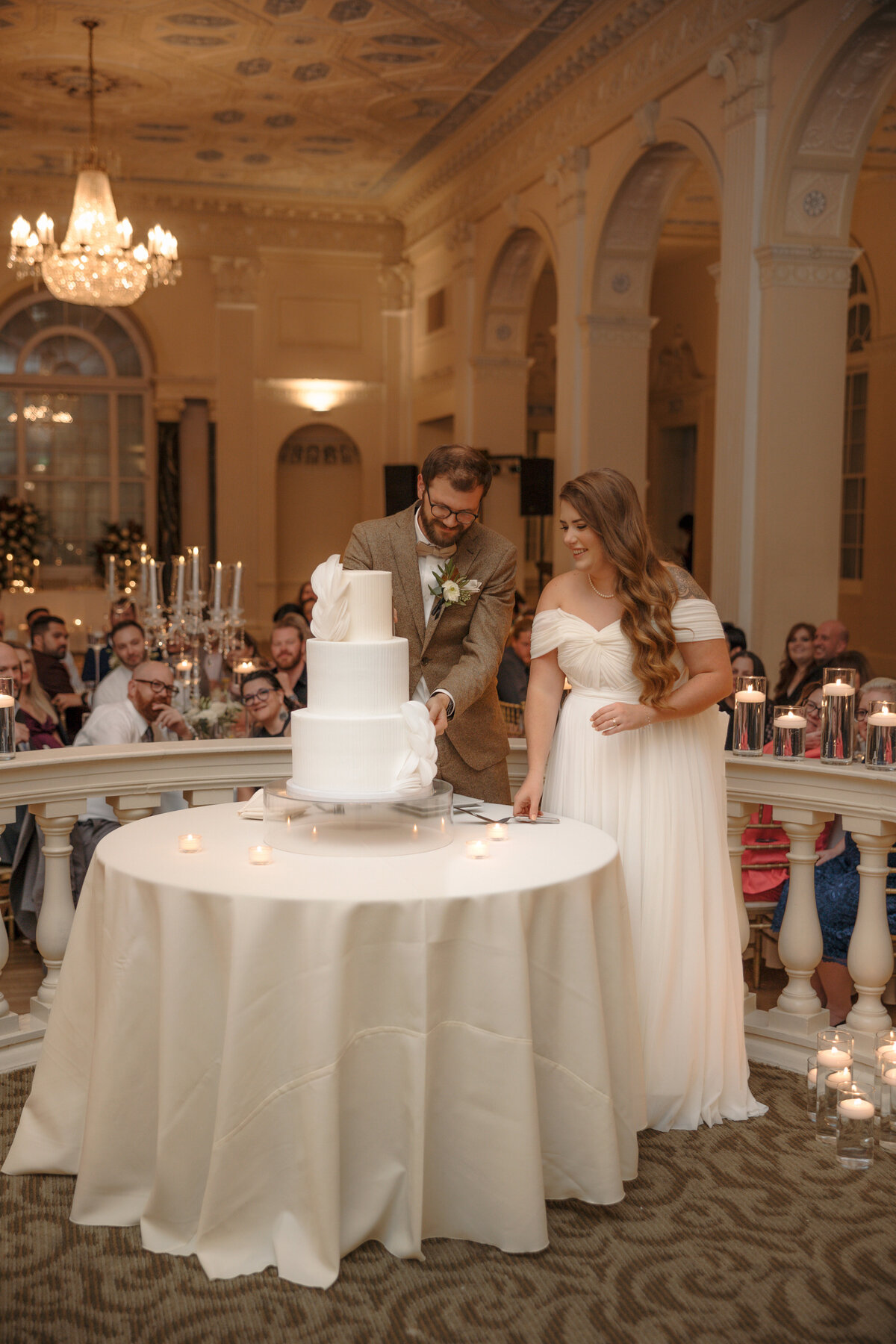 Bride and groom cutting their white tiered wedding cake in a candlelit ballroom.