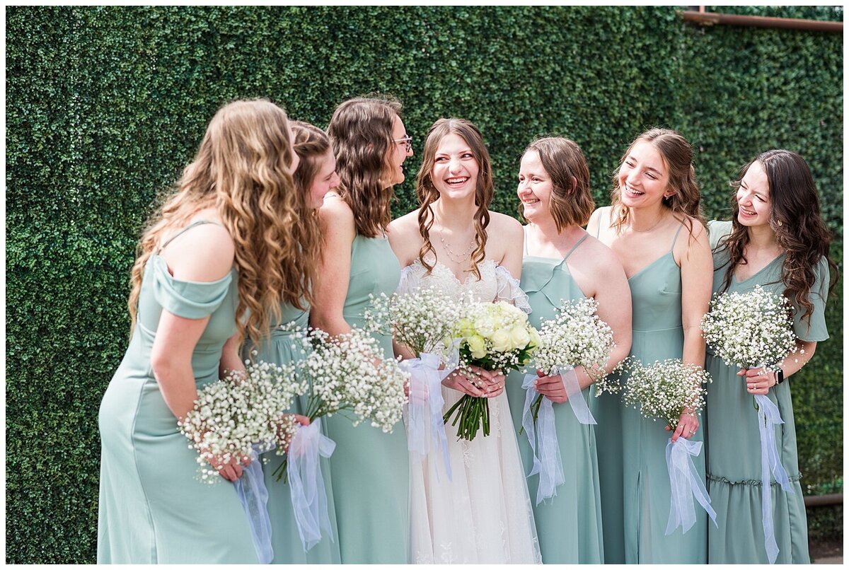 Bride stands between her 6 bridesmaids who are in green satin dresses holding their bouquets and they all look towards each other for a candid look