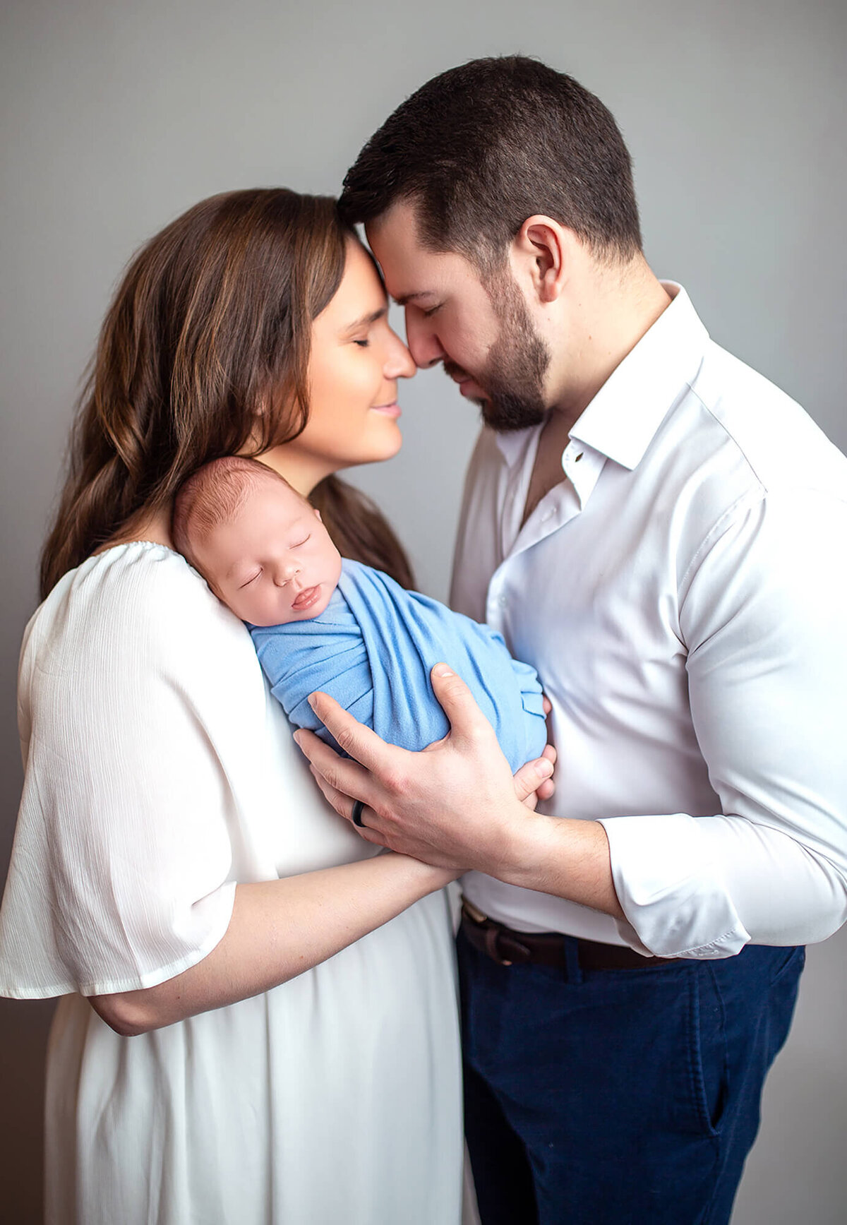 Mother and father standing forehead to forehead with newborn baby nestled on mothers shoulder wrapped in blue 
