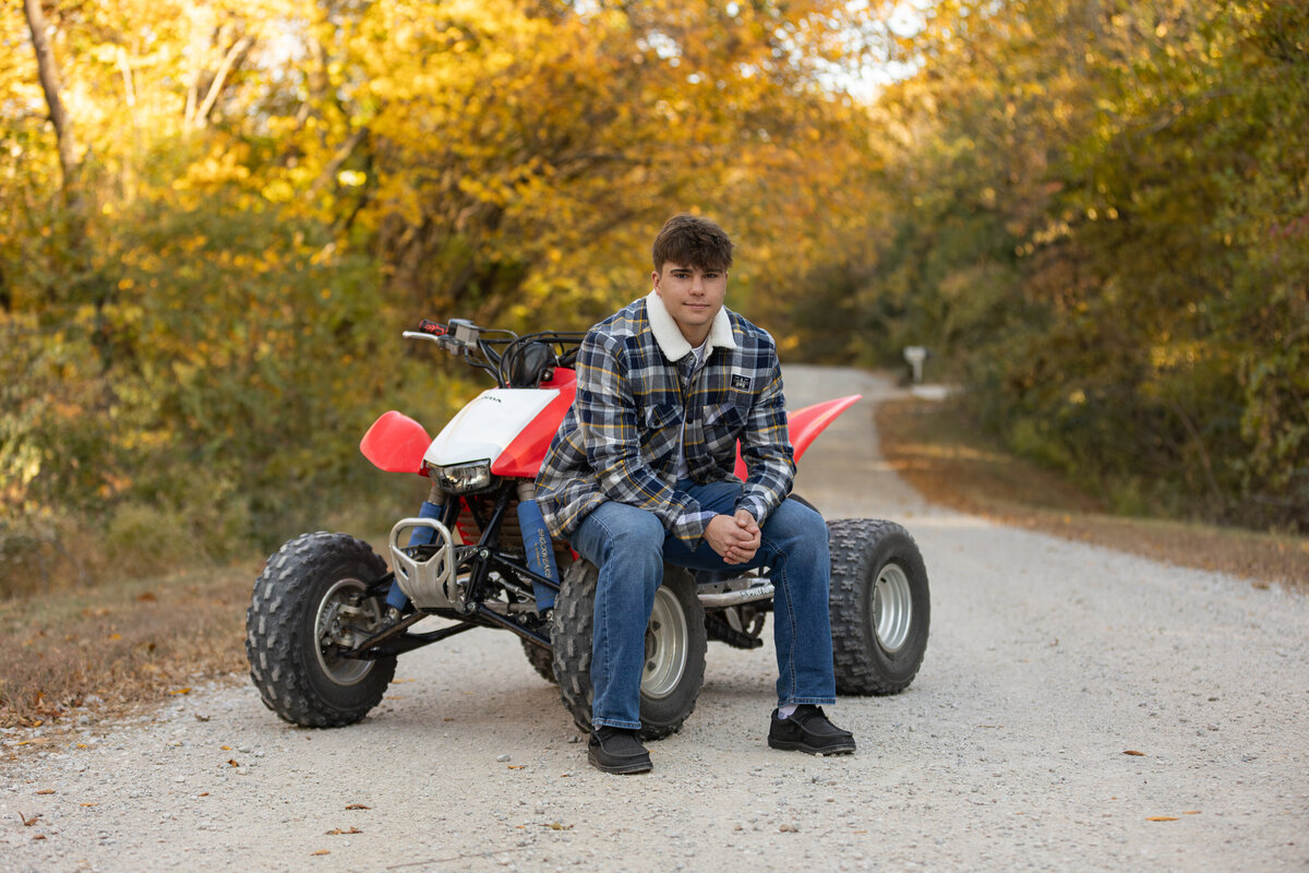 A high school senior boy sits on a red and white ATV along a scenic dirt road lined with vibrant autumn foliage. Wearing a plaid sherpa-lined jacket and jeans, he exudes a laid-back and adventurous vibe. This rustic outdoor portrait perfectly captures his personality and love for the outdoors.