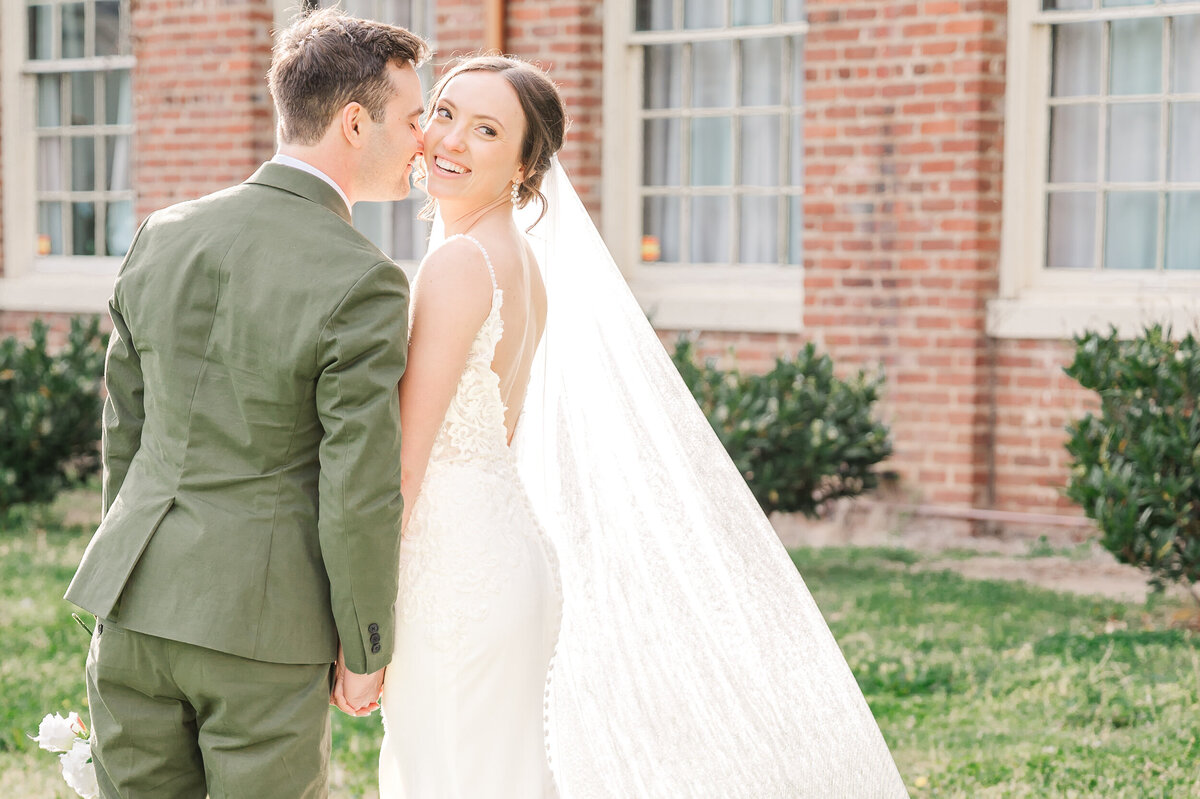 A stunning bride getting kisses by her husband in a green tux on her temple next to a brick wall with greenery on their wedding day in Virginia as they enjoy their Raleigh wedding photos by JoLynn Photography