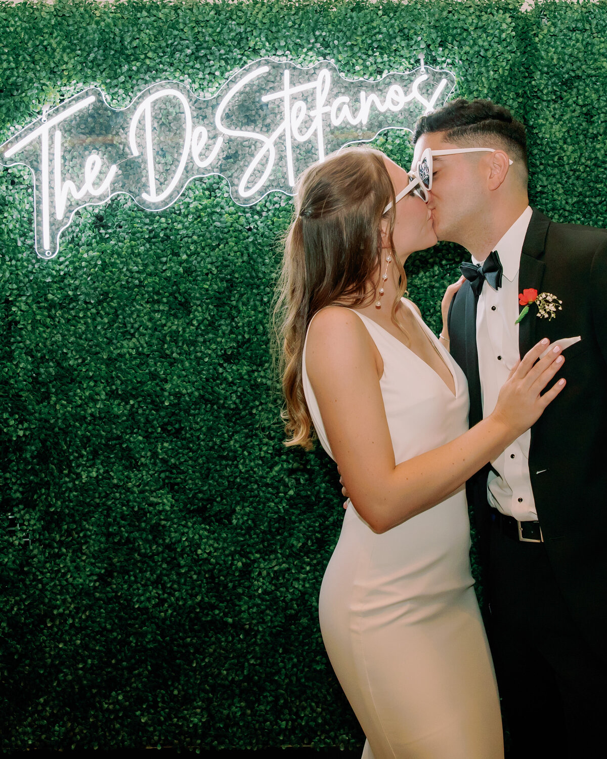A wedding couple kissing in front of a neon sign that reads "The De Stefanos"