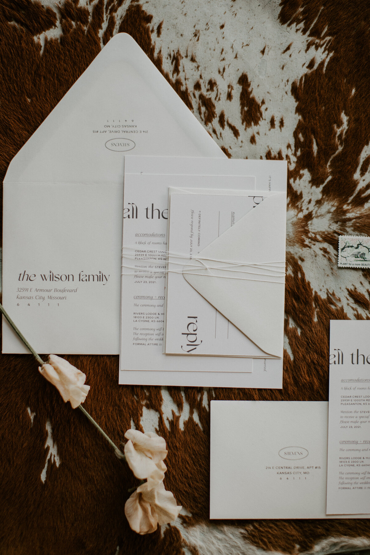 Various white wedding stationery with brown font  and flower atop brown and white cow hide.