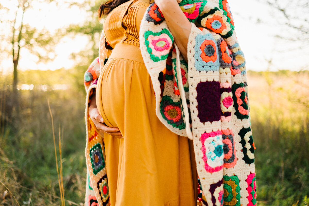 Beautiful belly photo of a pregnant woman in a garden, showcasing her radiant glow. She wears a long yellow dress paired with a colorful square wool jacket, capturing a vibrant and serene maternity moment.