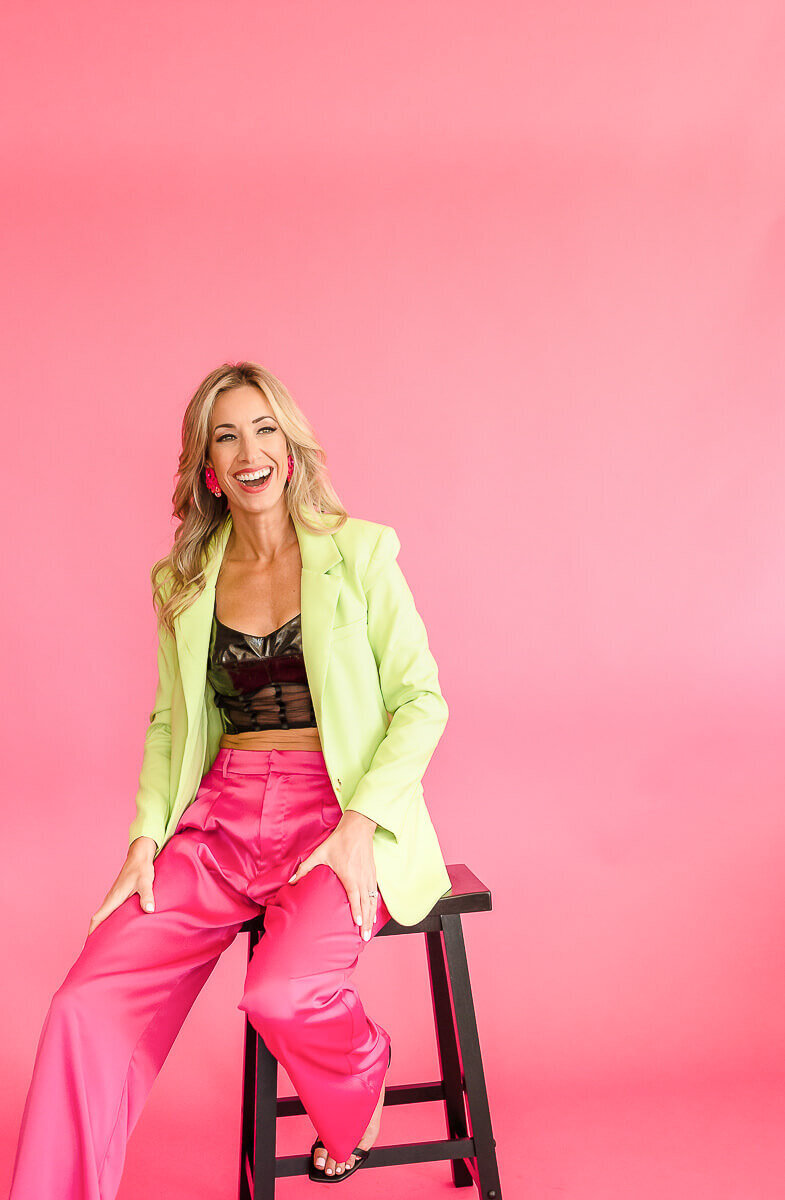 Woman in pink pants and lime green blazer sitting on stool with pink background.