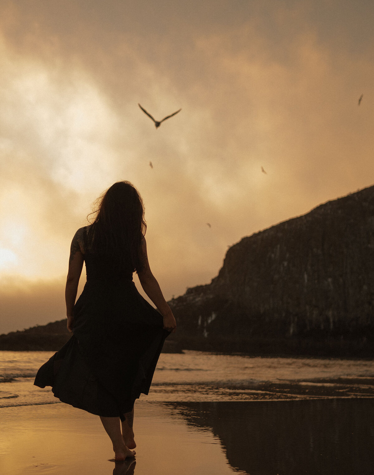 woman walks along Oregon Coast shoreline in golden light as the seagulls fly around her