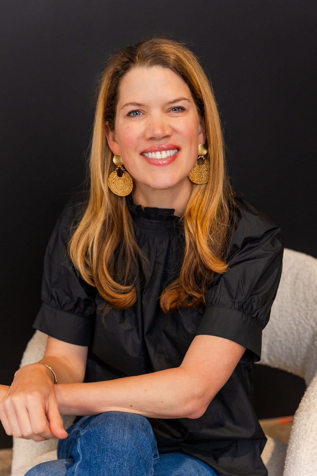 woman wearing a black top and golden earrings sitting sideway hands together on a chair in front of a black background during portraits in Atlanta studio with Laure Photography