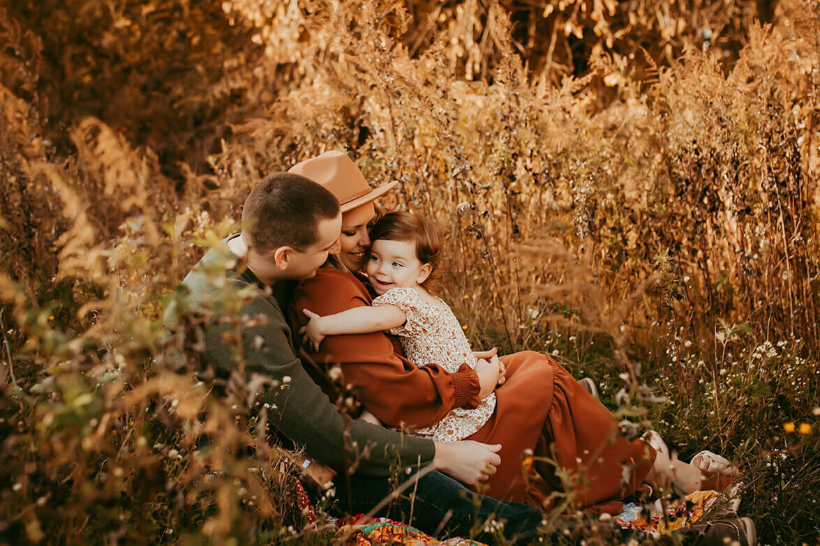 a two year old girl hugging her mom who is sitting in dads lap in a field
