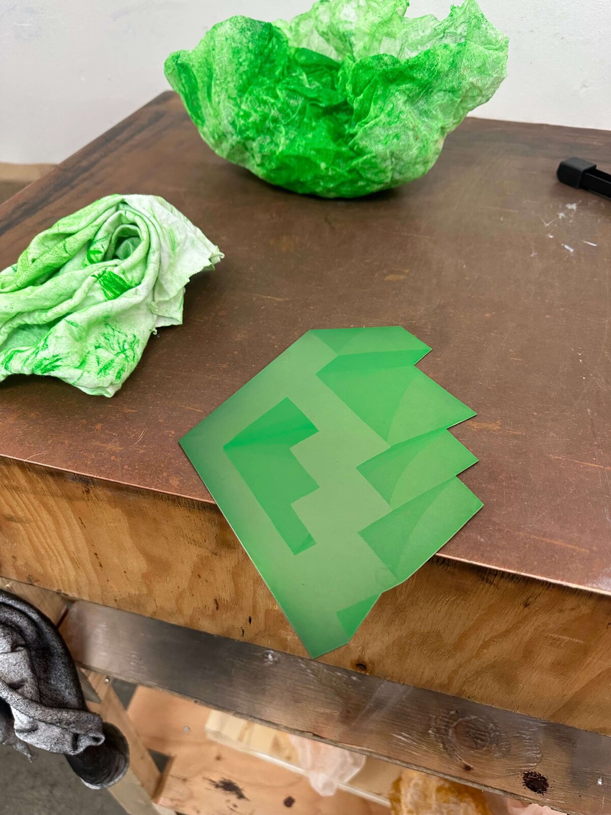 A green copper plate showing tonal steps from an aquatint etch, resting on a wooden table with green-stained cloths nearby.