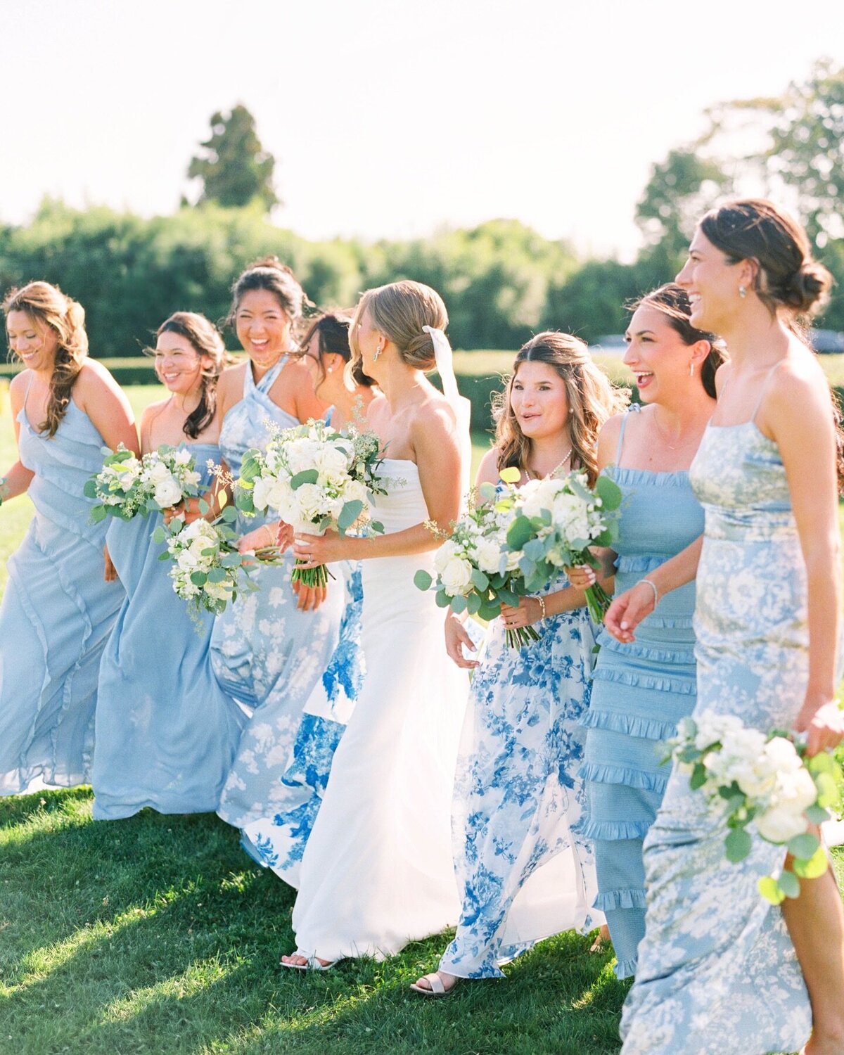 Wedding Photographer RI | A bride in a white gown walks joyfully with bridesmaids in different light blue dresses, holding bouquets. The group is outdoors on a sunny day.