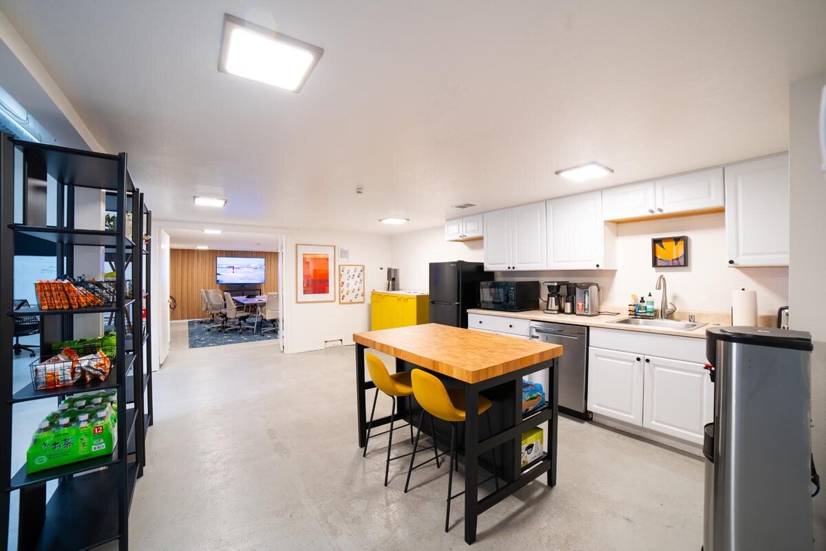 Breakroom kitchen with white cabinetry, a butcher-block island, yellow stools, and open shelving stocked with snacks.