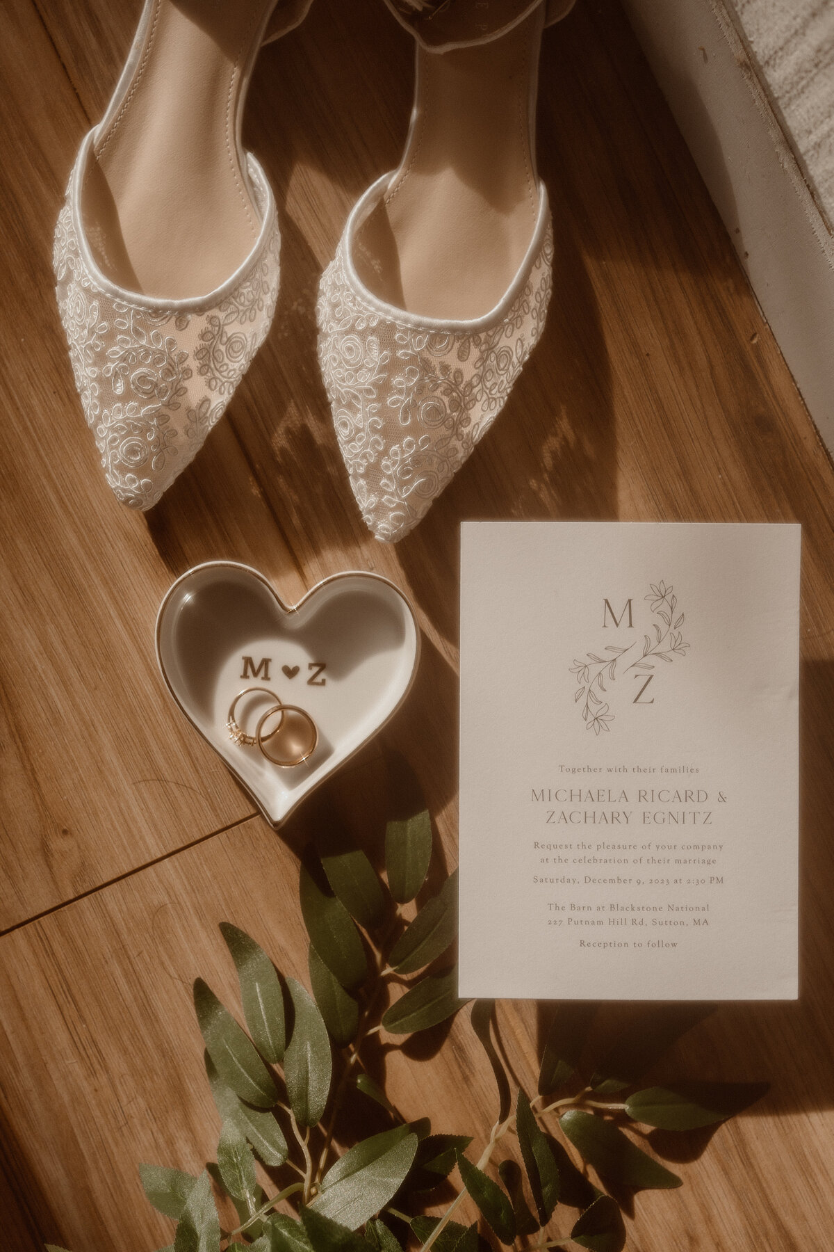 Elegant flat lay of wedding details featuring lace bridal shoes, wedding invitation, gold wedding rings in a heart-shaped dish, and green foliage. Ideal for couples looking for a documentary-style wedding photographer to capture artistic and detailed shots of their special day.