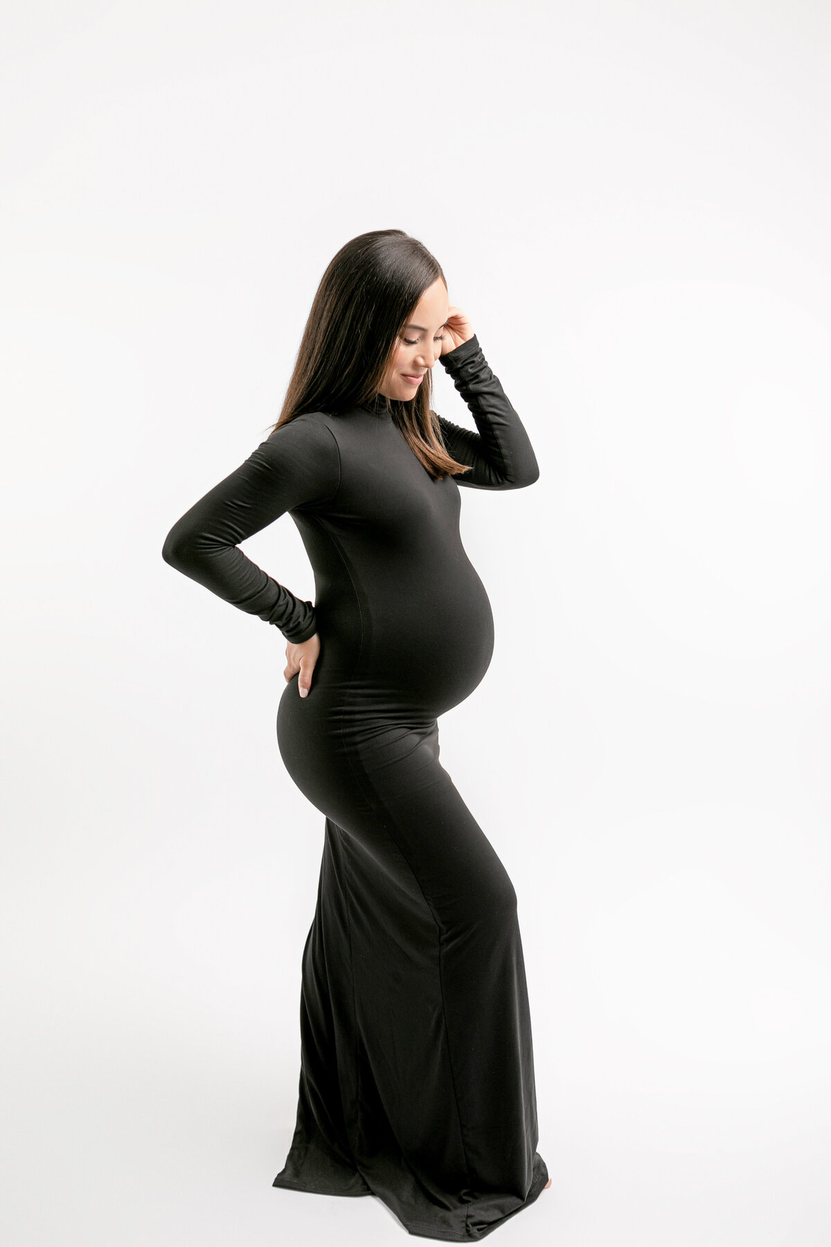 Expectant mother, wearing a fitted black dress in an all white studio looking at her pregnant belly with fresno, ca maternity photographer
