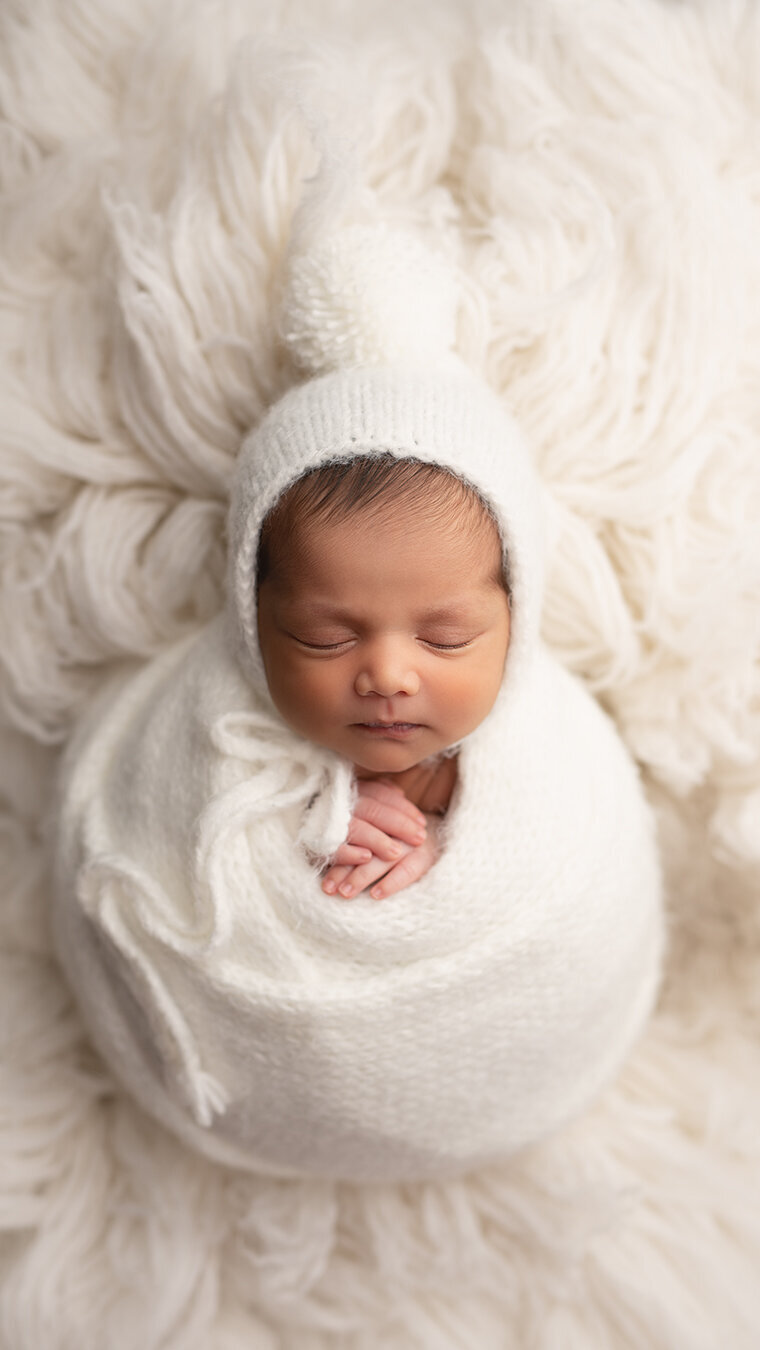 Newborn baby peacefully sleeping in a white knit wrap and bonnet surrounded by soft white textures — photographed by Meraki Photographic Creations in Paris, Ontario for the Newborn Investment & Session Pricing mobile page.