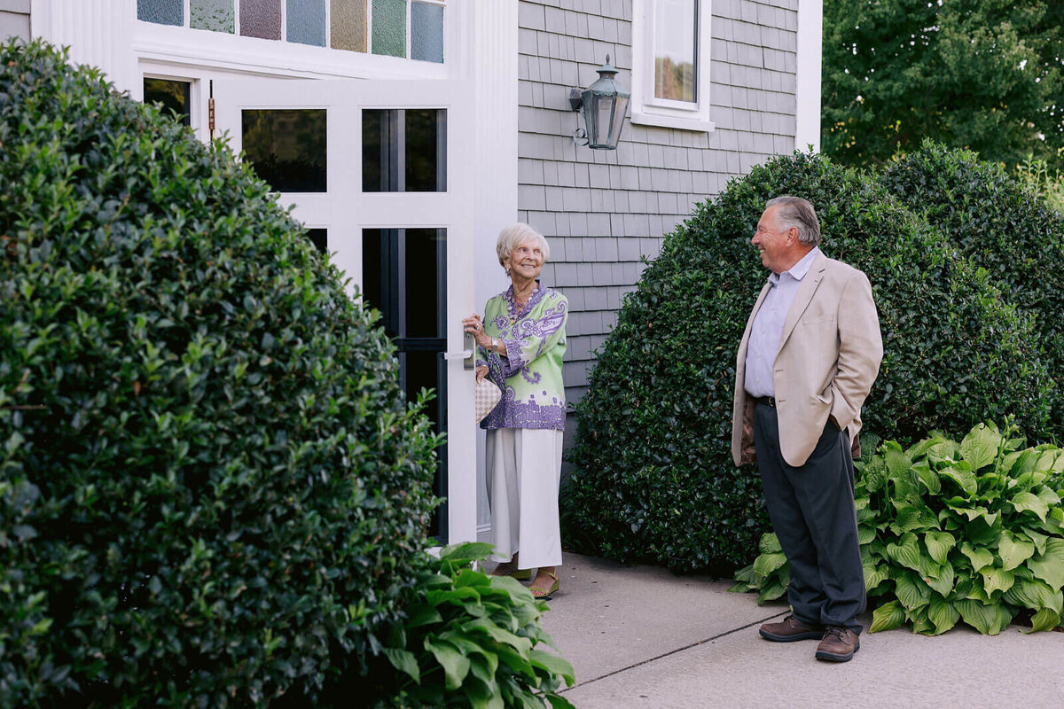 An old, woman holding the door, and an old man are smiling at each other outside a house at Cape Cod, Osterville, MA.