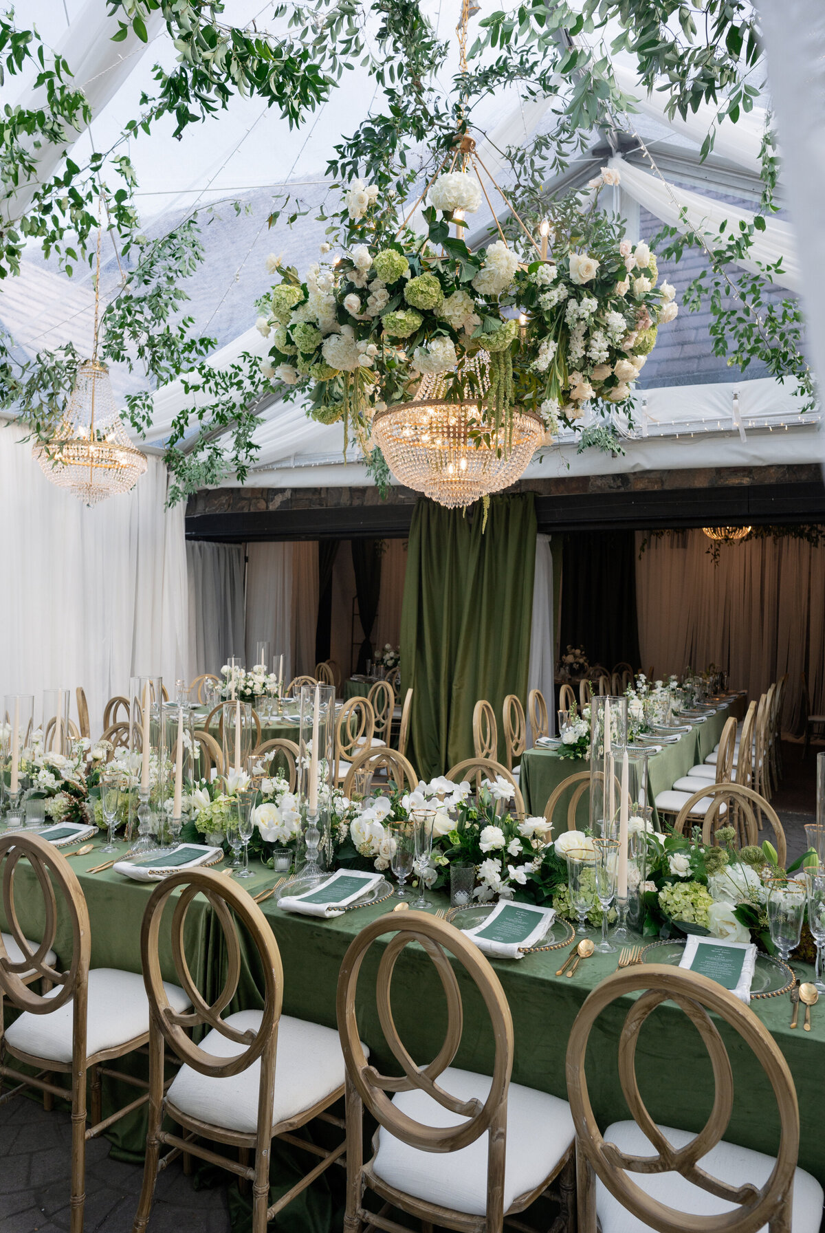 Luxurious reception tablescape with green linens, white florals, and hanging floral chandeliers inside a tented wedding reception.