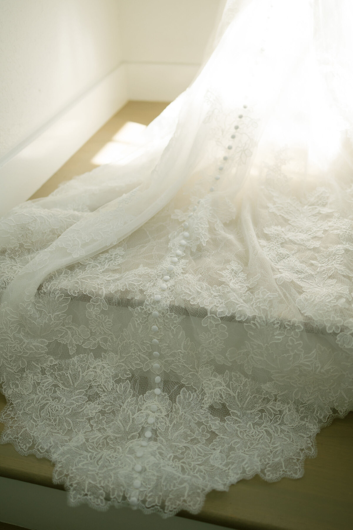 Close-up of an intricate lace wedding gown train showcasing delicate floral embroidery and a line of fabric-covered buttons, photographed in soft natural light for romantic bridal detail inspiration.