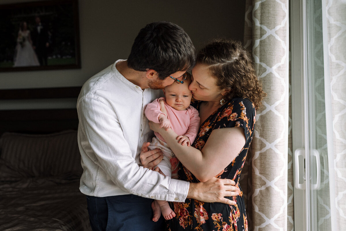 mom and dad holding and kissing baby wearing pink next to window