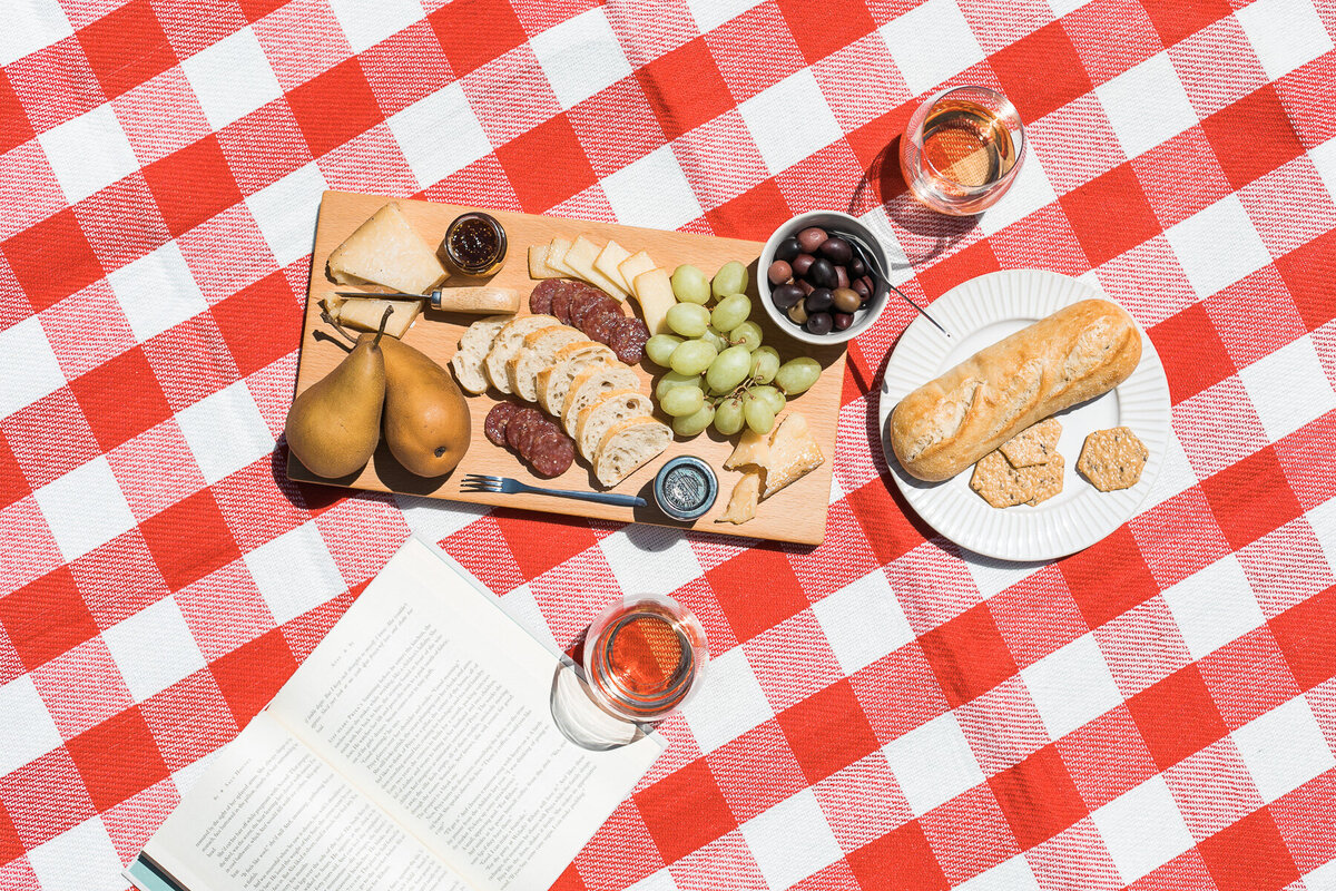 wine and fruit and bread charcuterie on a red checkered picnic blanket with a book
