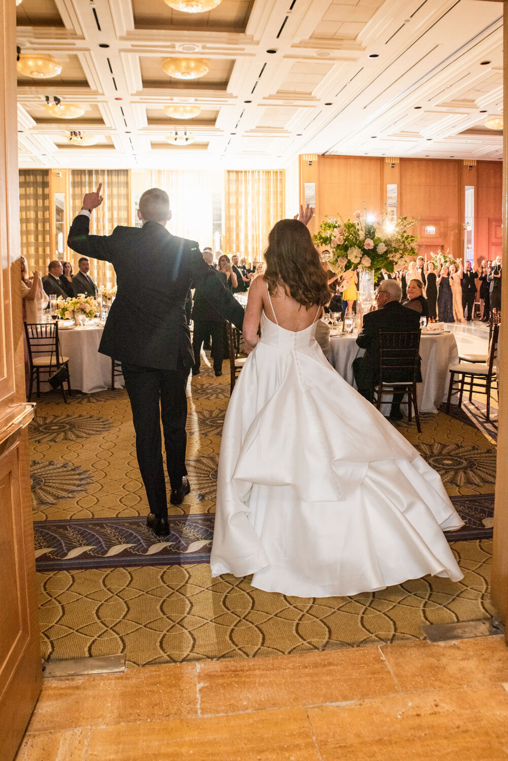 A bride and groom are walking into a room with tables and chairs