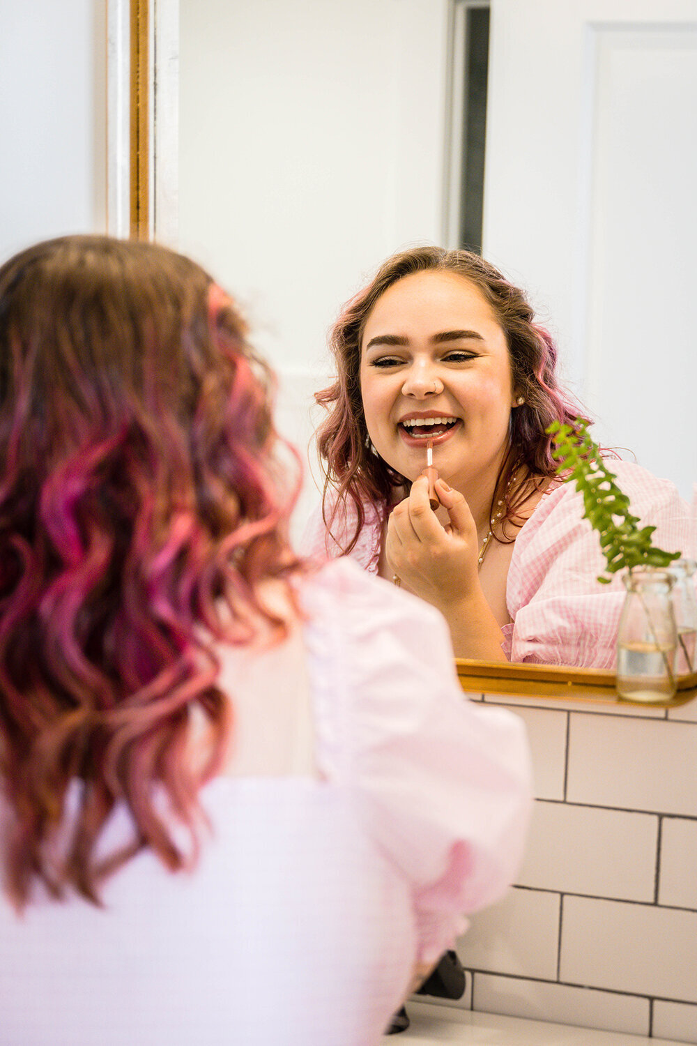 A marrier on her elopement day gets up and close in the mirror as she gets ready. She is smiling as she applies lip gloss in a clean and modern hotel bathroom.