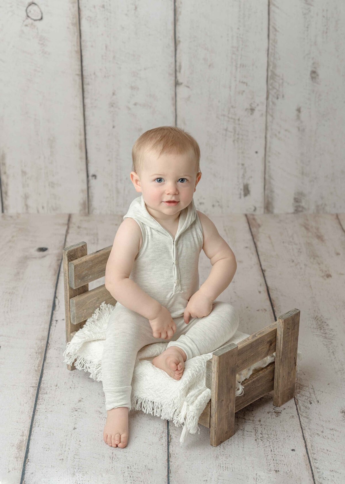 One year old boy sitting on small wood bed, dressed in oatmeal colored outfit