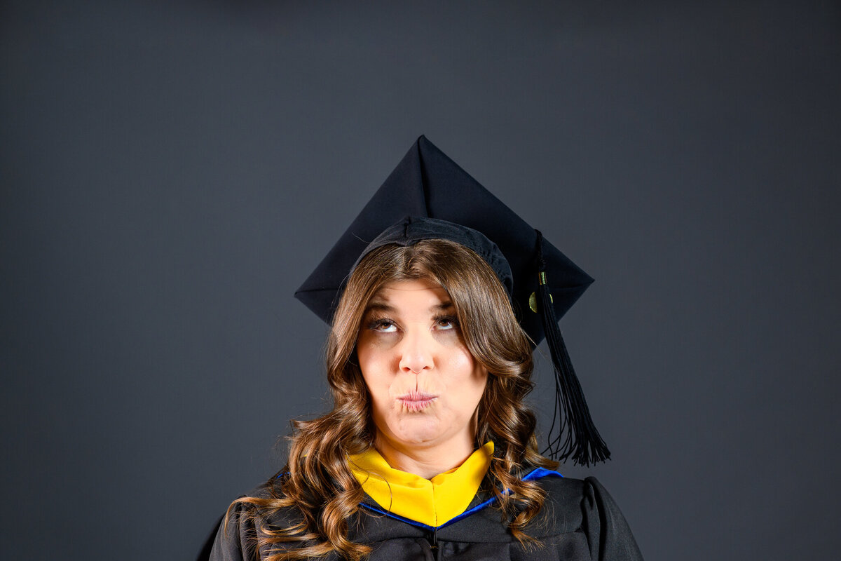 A girl dressed in a graduation robe and cap makes a funny face at the camera, with a simple gray background behind her