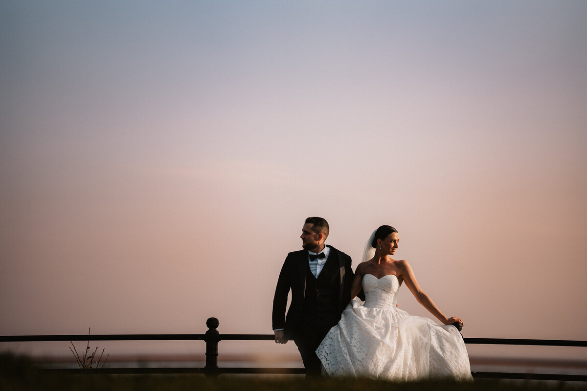 Bride and groom posing by the seaside railings at sunset at a luxury Lytham wedding venue, with soft pink skies and elegant wedding attire.