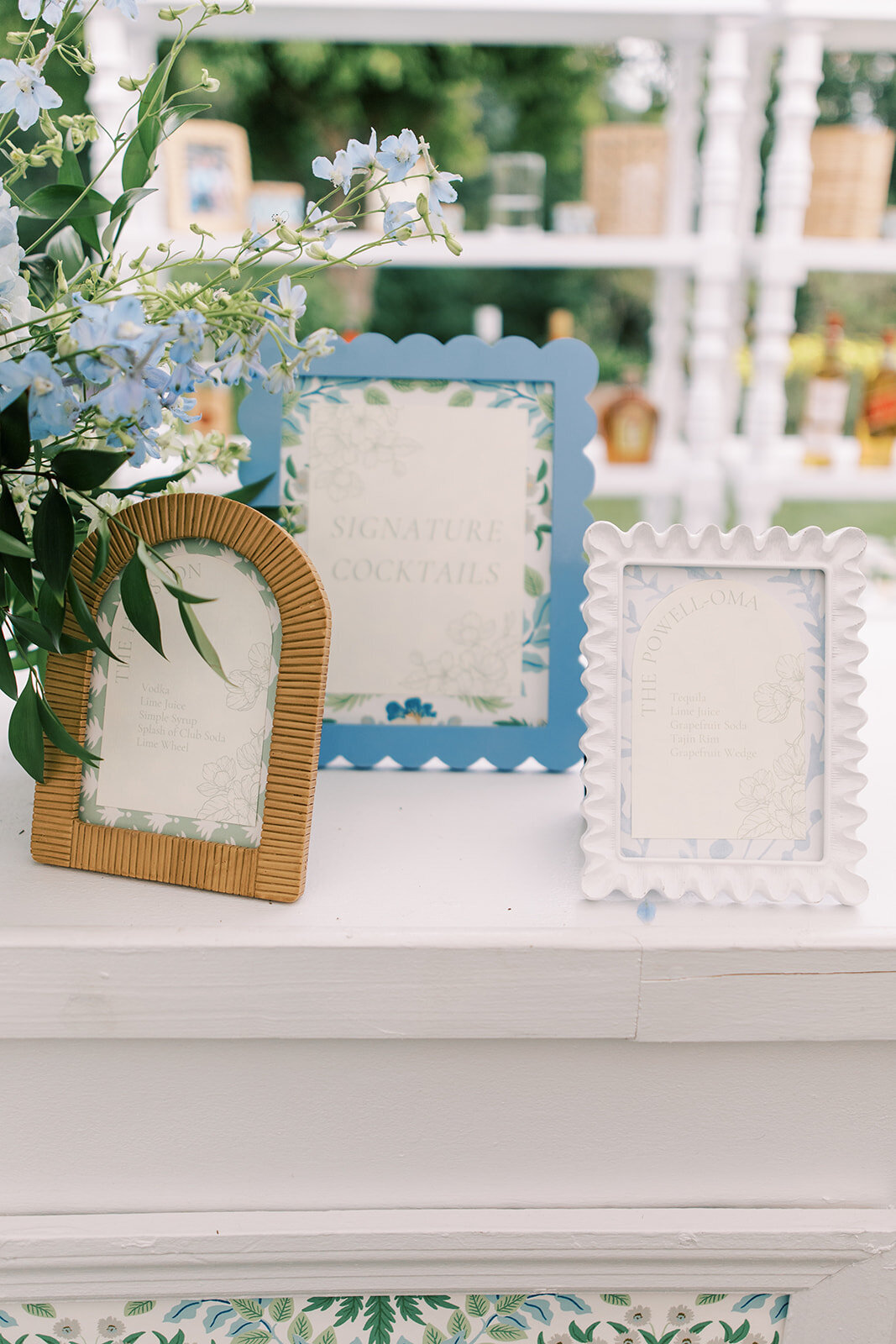 Three framed bar menus on a white bar sitting next to elegant blue and white florals. 