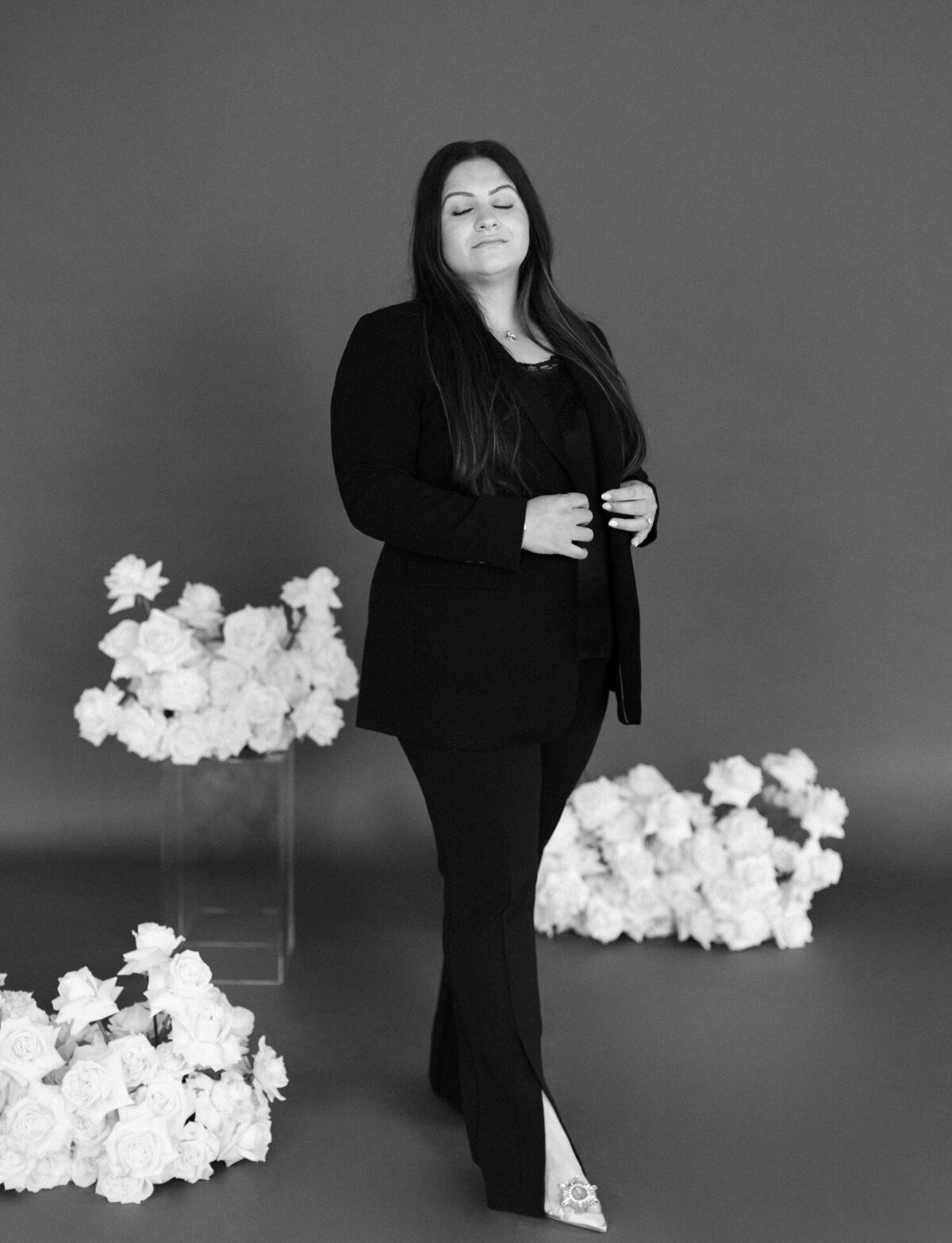woman in black pants and black blazer looks peaceful among white roses, dark background for florist brand photos