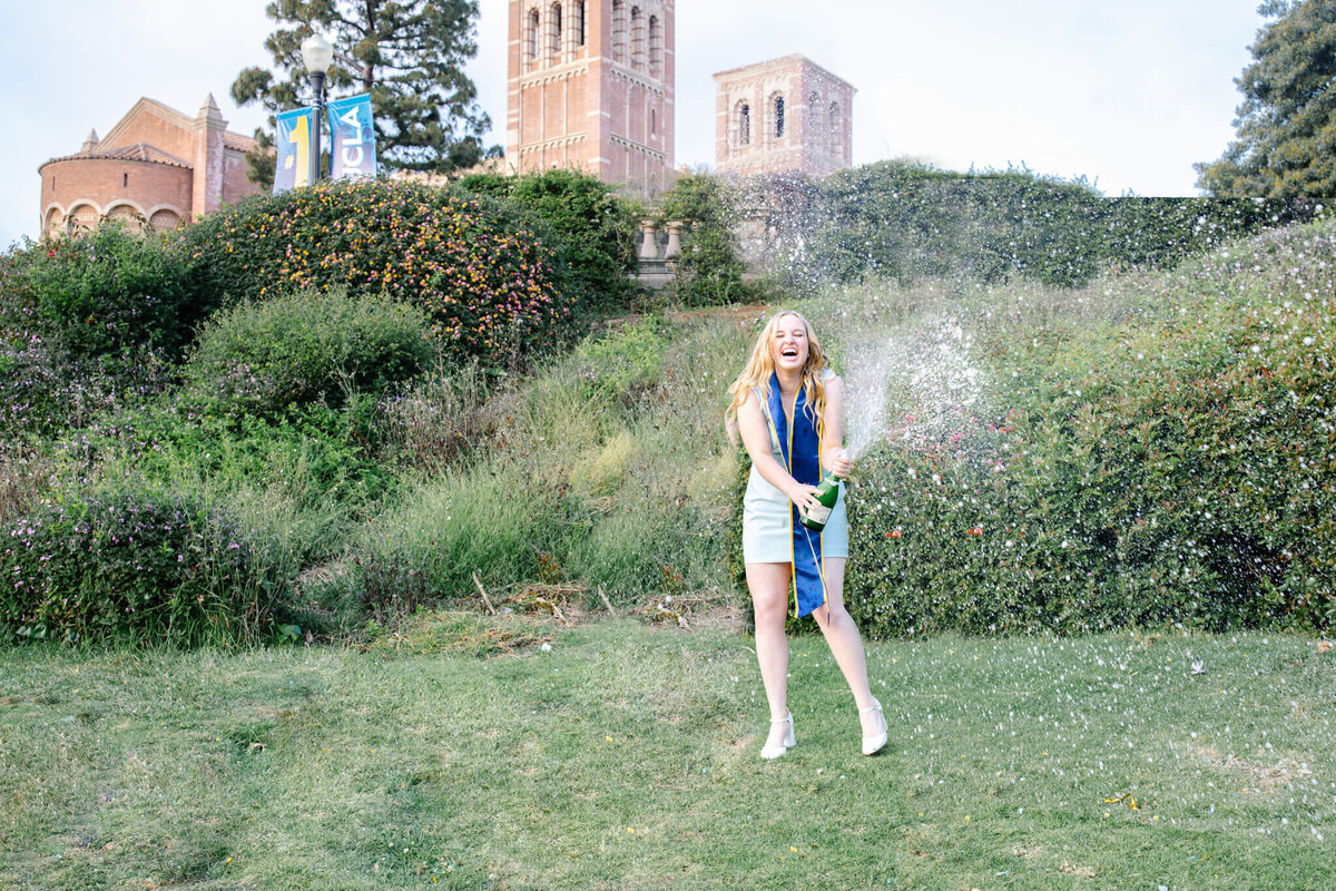 Blonde UCLA graduate in a light blue dress popping a bottle of champagne on a hill in front of Royce Hall